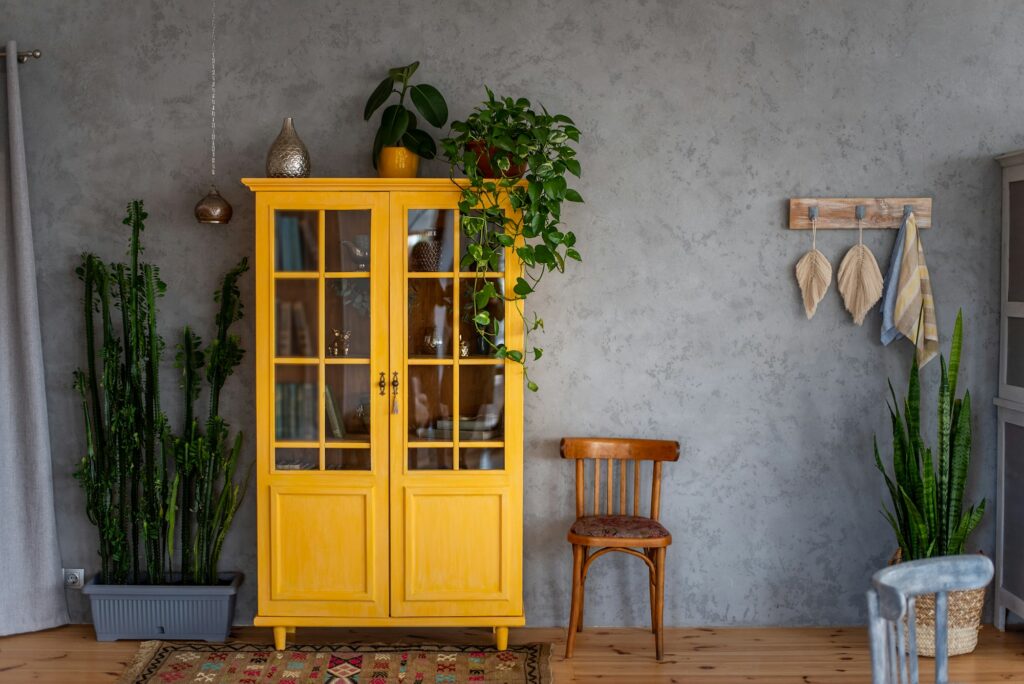 Deep yellow wooden bookcase against gray concrete wall. Around green pots with home flowers.