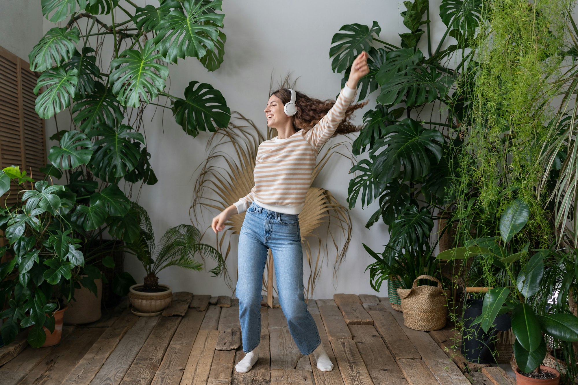 Happy woman wear wireless headphones listening to music, dancing in home garden with tropical plants