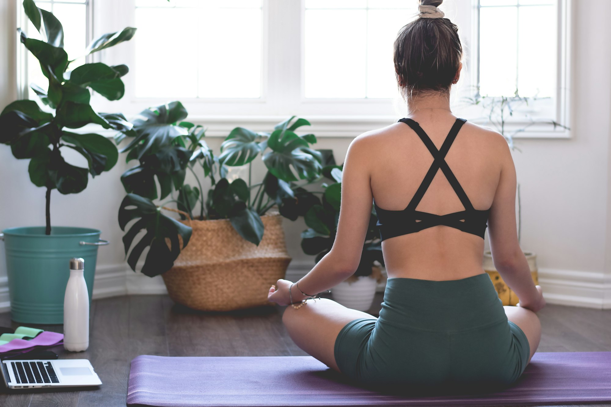 Young girl woman sitting in yoga meditation pose facing large bright window and tropical houseplants